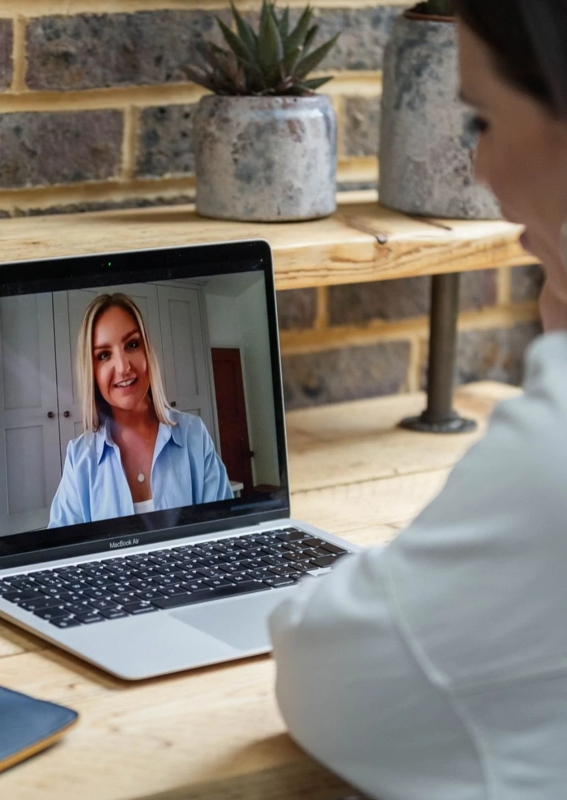 Person on a video call with a woman on a laptop screen, in a cozy indoor setting with brick walls and wooden shelves with potted plants.