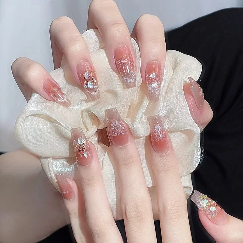 A close-up of a person's hands with decorated acrylic nails, holding a satin fabric bow. The nails are long, with a clear base, rhinestones, glitter, and white designs.