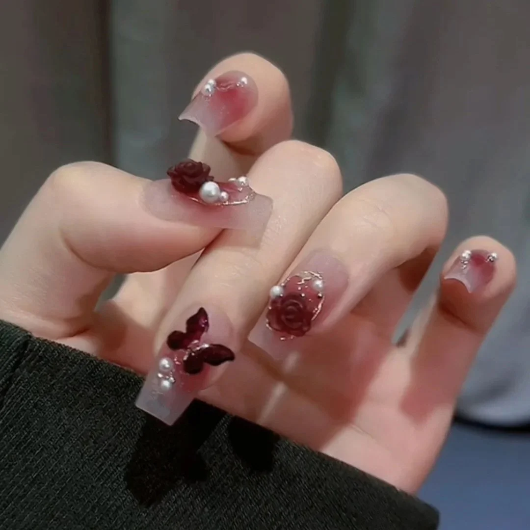 Close-up of a hand with decorated nails featuring dark red roses, small pearls, and shiny embellishments on clear acrylic nails.