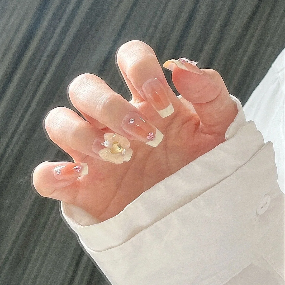 Close-up of a hand showing manicured nails with intricate nail art, including floral designs and 3D embellishments, with part of a white shirt cuff visible.
