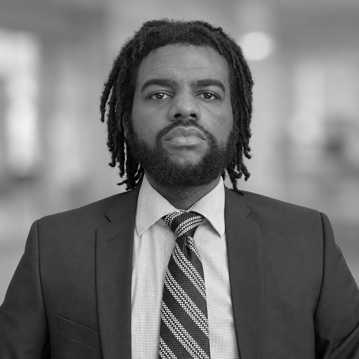A black and white portrait of a man with dreadlocks, a beard, wearing a suit, shirt, and tie, against a plain background.