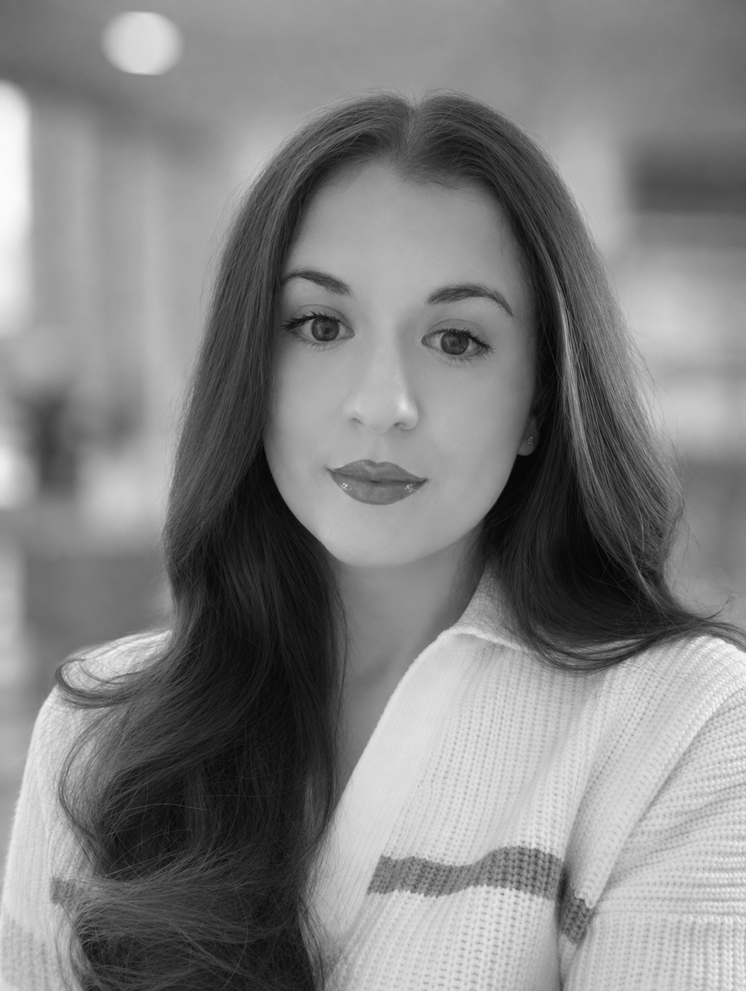 A black and white portrait of a person with long hair, wearing a sweater, indoors.