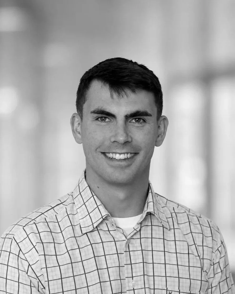 Black and white portrait of a young man smiling, wearing a checkered shirt, with a blurred background.