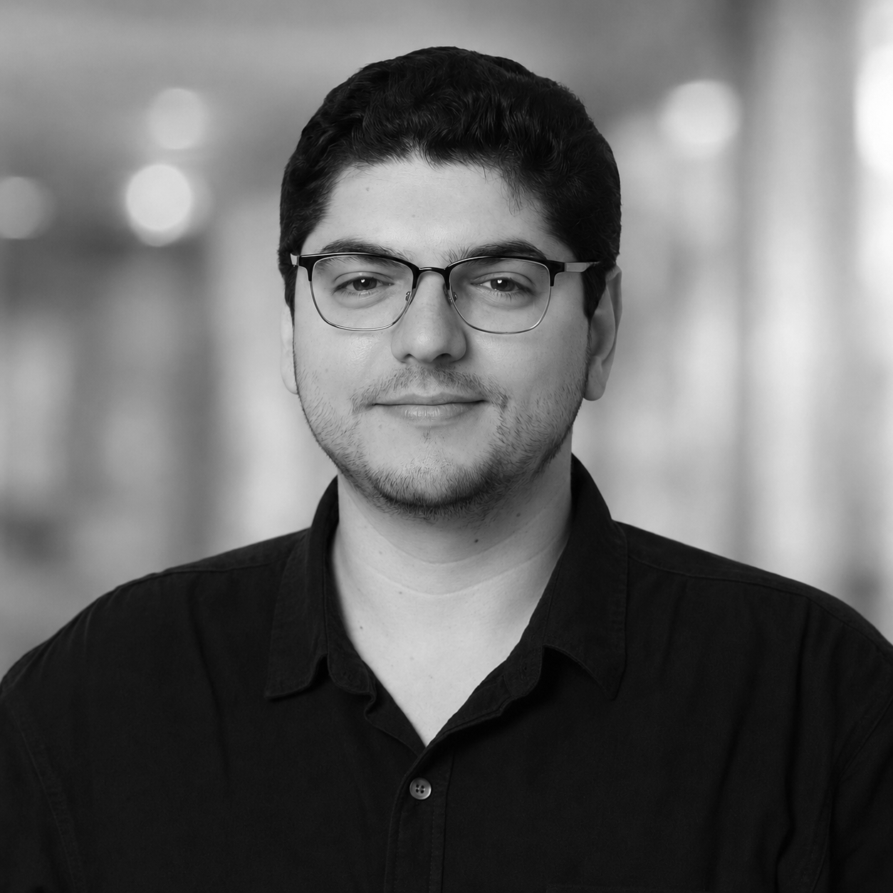 Black-and-white professional portrait of a young man with glasses, short dark hair, wearing a dark shirt, smiling slightly, against a plain background.