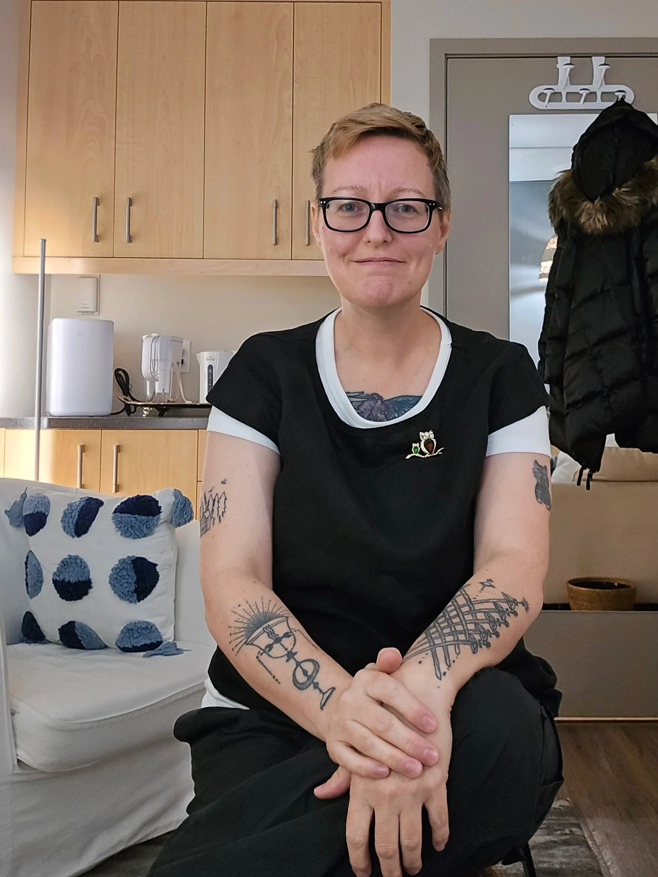 Registered clinical counsellor Melanie Armstrong, a white women with short blonde hair wearing a black and white shirt and sitting on a chair in a small office space with a kitchenette and white couch.