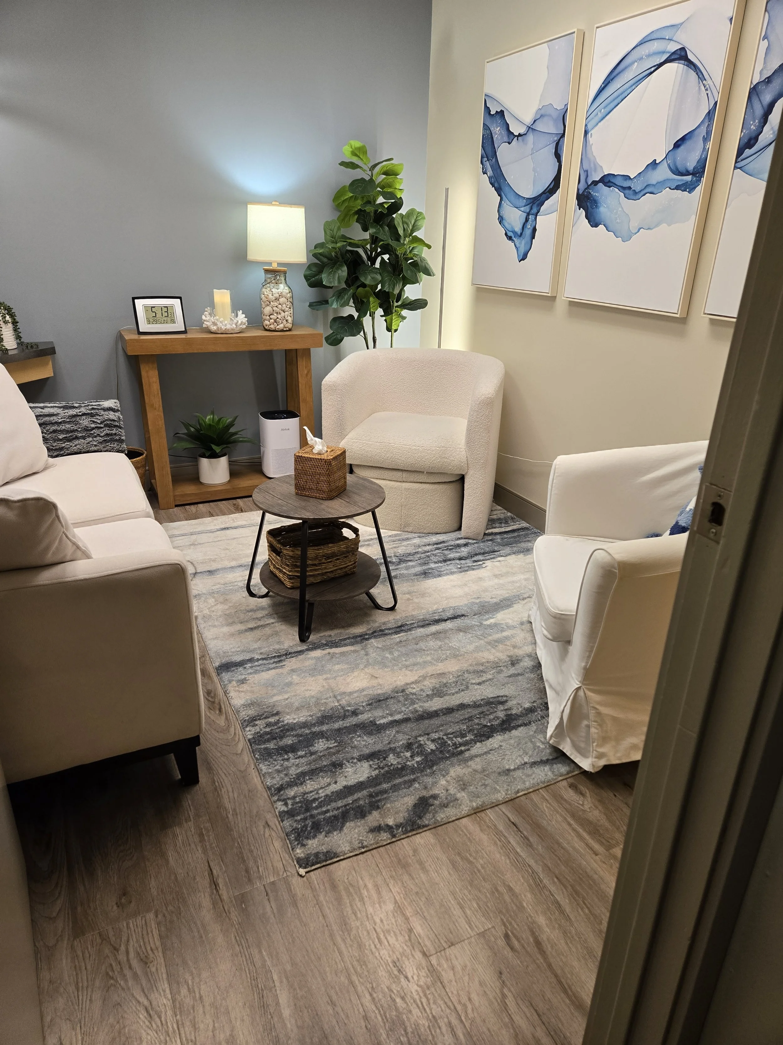 A counselling space with blue walls, blue and white carpet, white chairs and a white couch, green plants, shelves, and three pictures on the wall of blue fluid swirling on a white background.