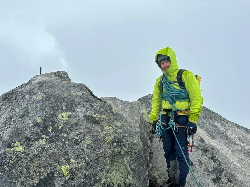 En person i fjellklatringutstyr står på en fjelltop og smiler, omgitt av store steiner og en grå himmel.