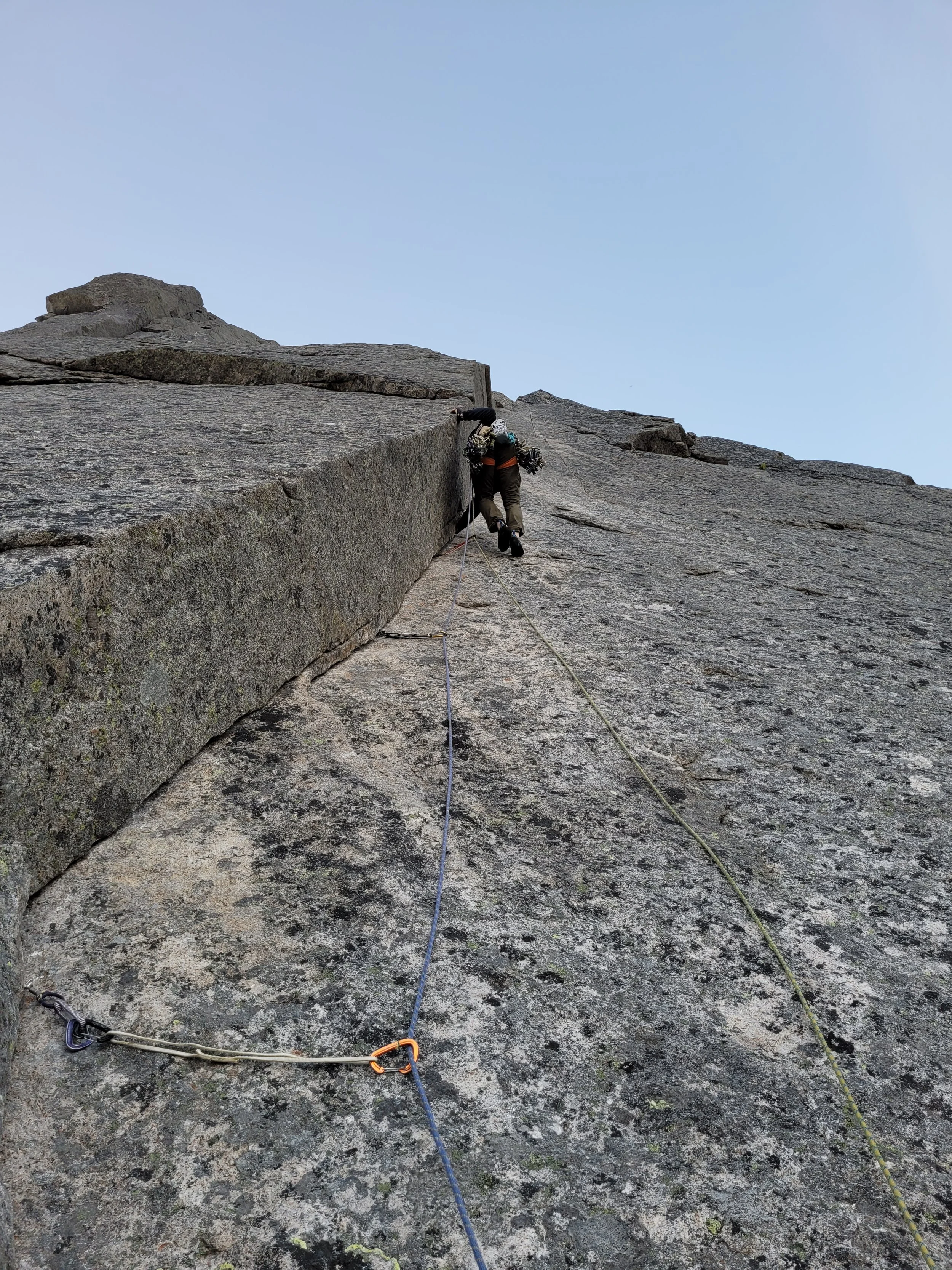 En klatrer opp en fjellvegg med tau og sikringer, iført klatretøy og hjelmer, mot en klar blå himmel.