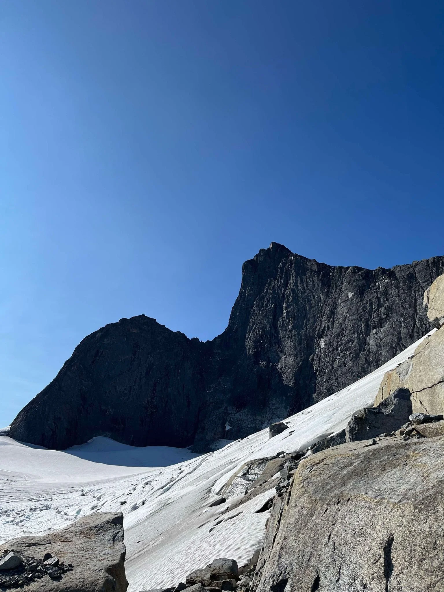 Snødekt fjellandskap med store steiner og en bratt fjellvegg under en klar blå himmel.