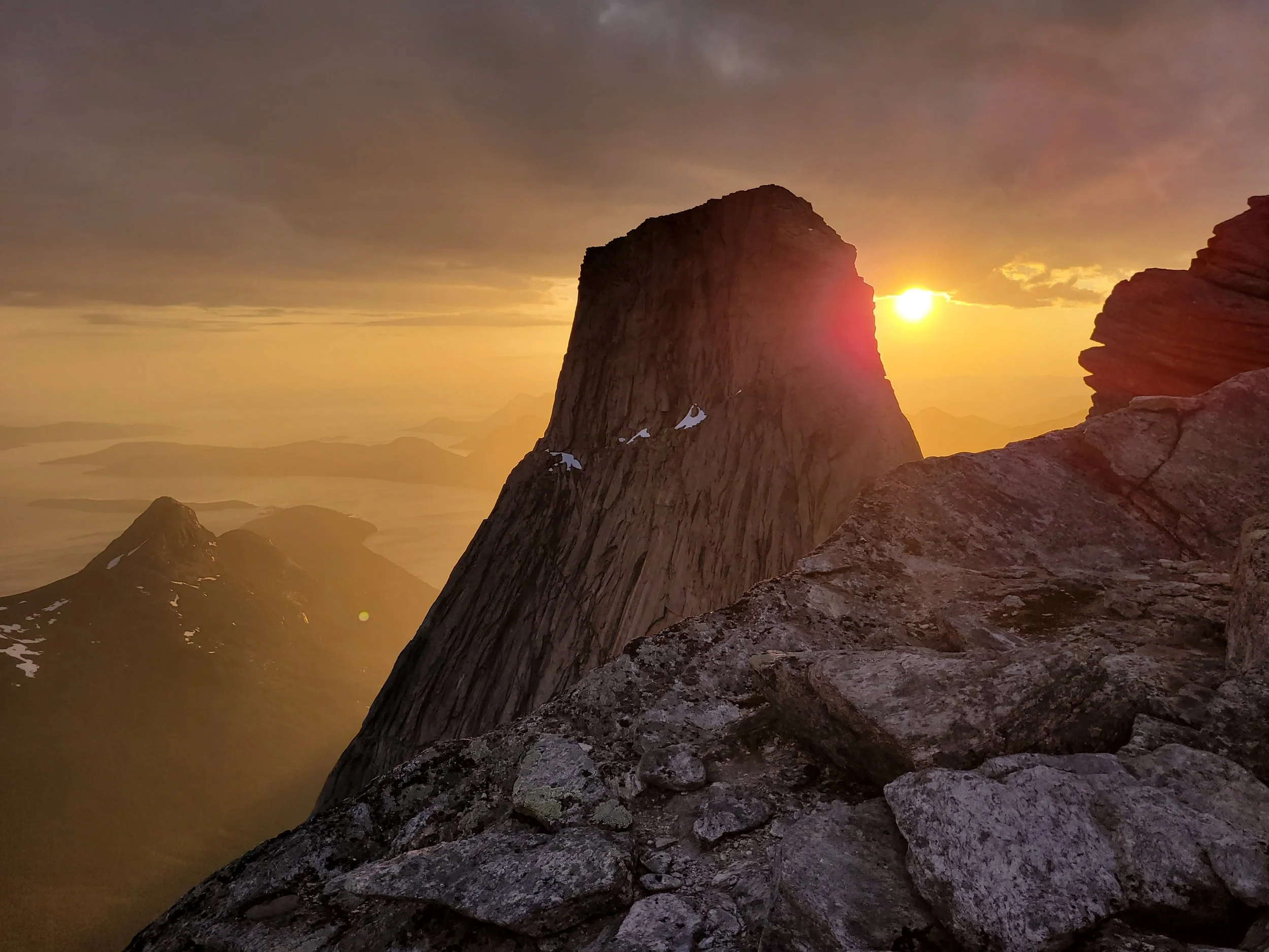 Et fjell med snøflekker på toppen under en solnedgang, sett fra en steinet fjellside.