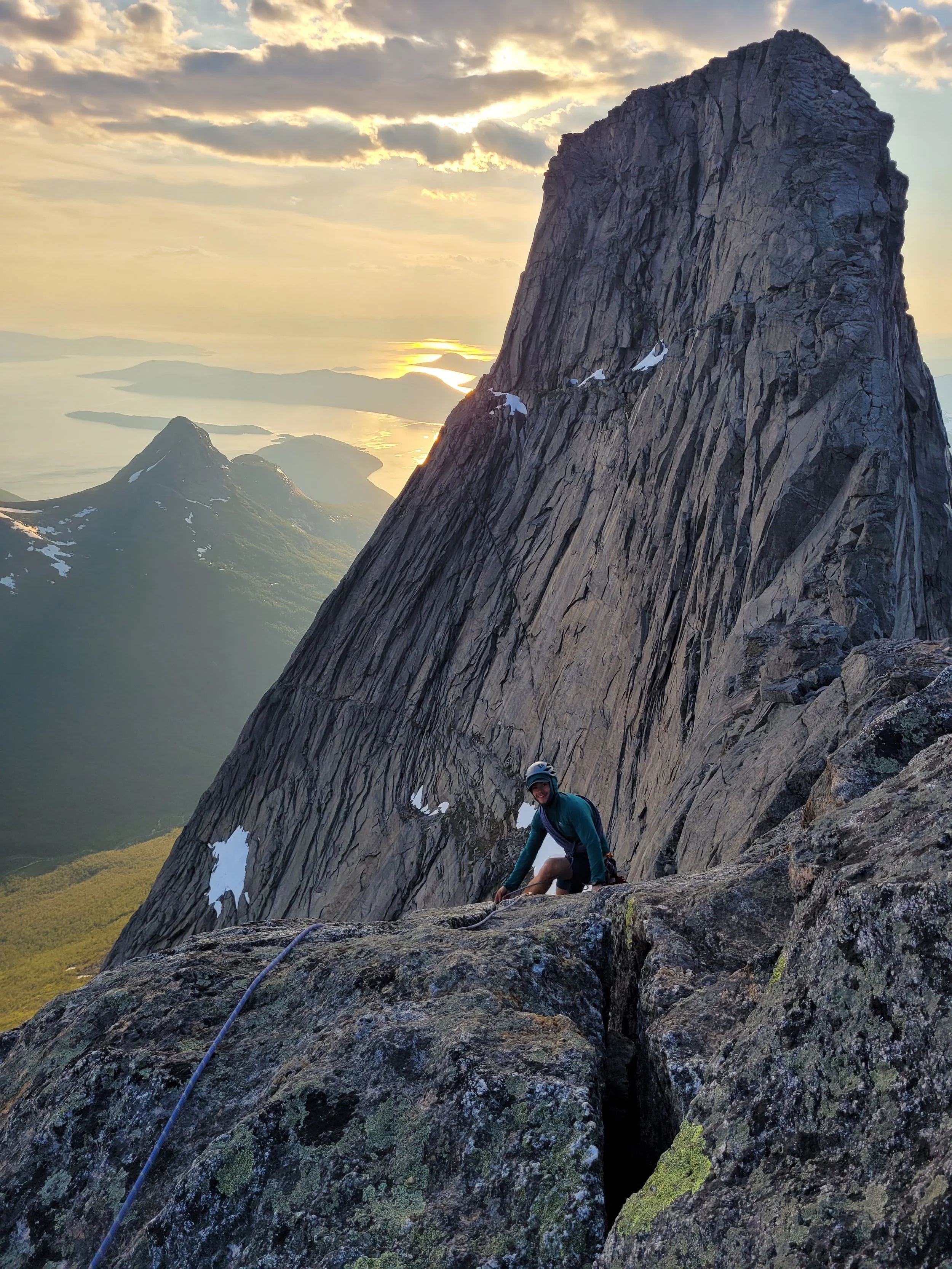 En person klatrer opp en bratt fjellvegg med fjell og innsjøer i bakgrunnen, under en solnedgang.