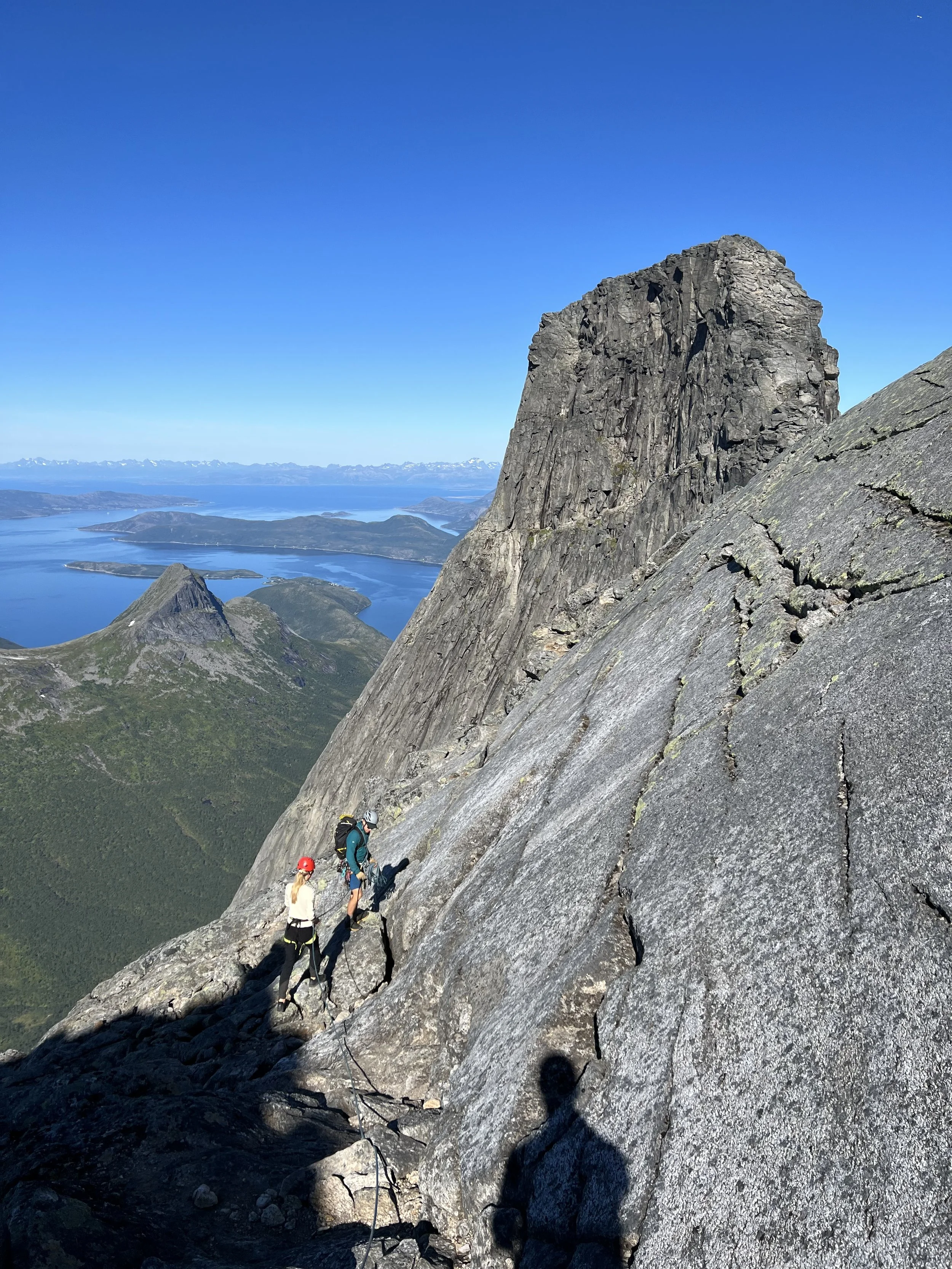 To klatrere i fjellsiden med tauer og hjelmer, omgitt av høye fjell med en innsjø og fjell i bakgrunnen under en klar blå himmel.