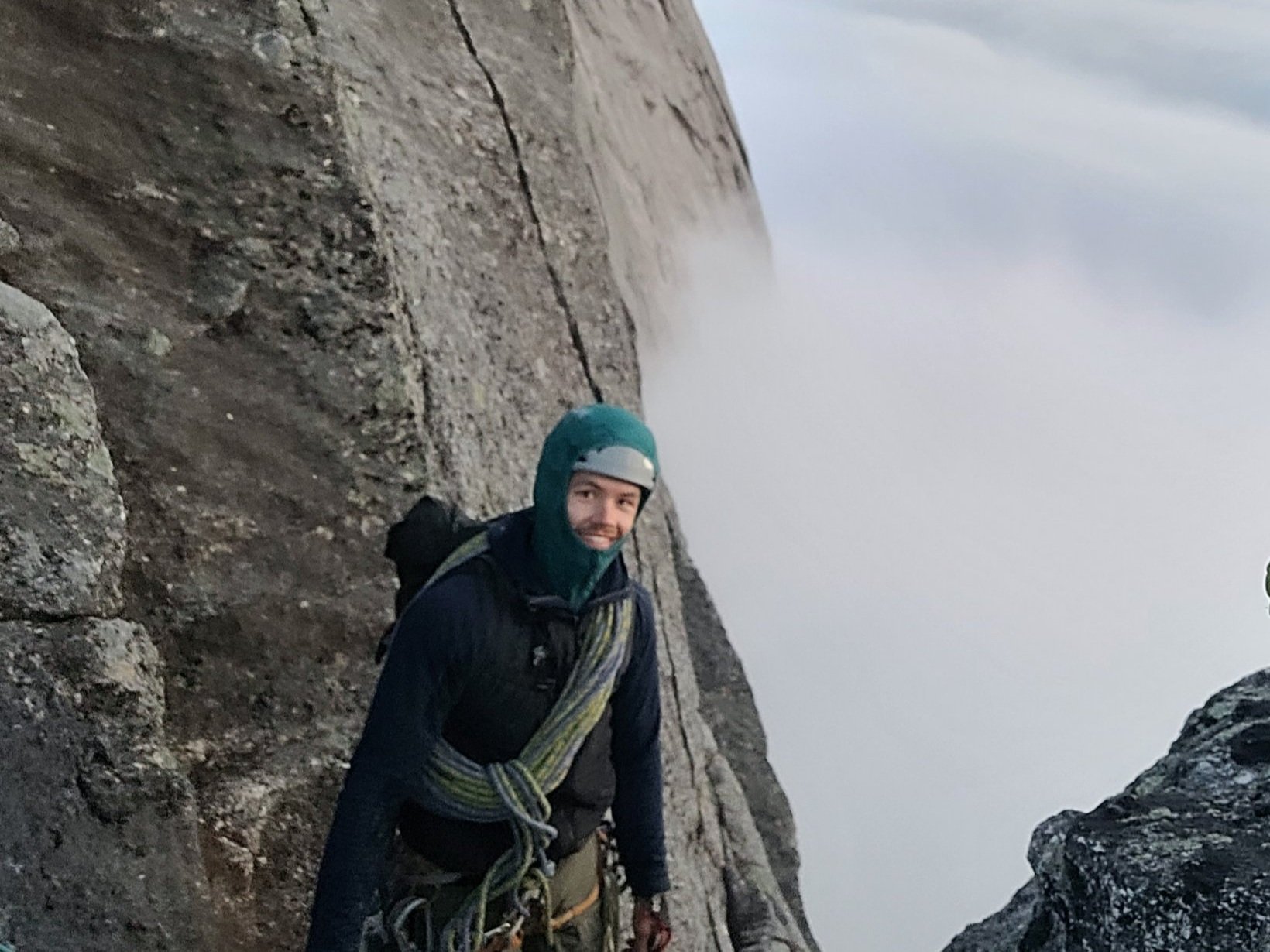 En person som klatrer på en fjellvegg i fjellet, omgitt av skyer og steiner, iført klatreutstyr og resekk.