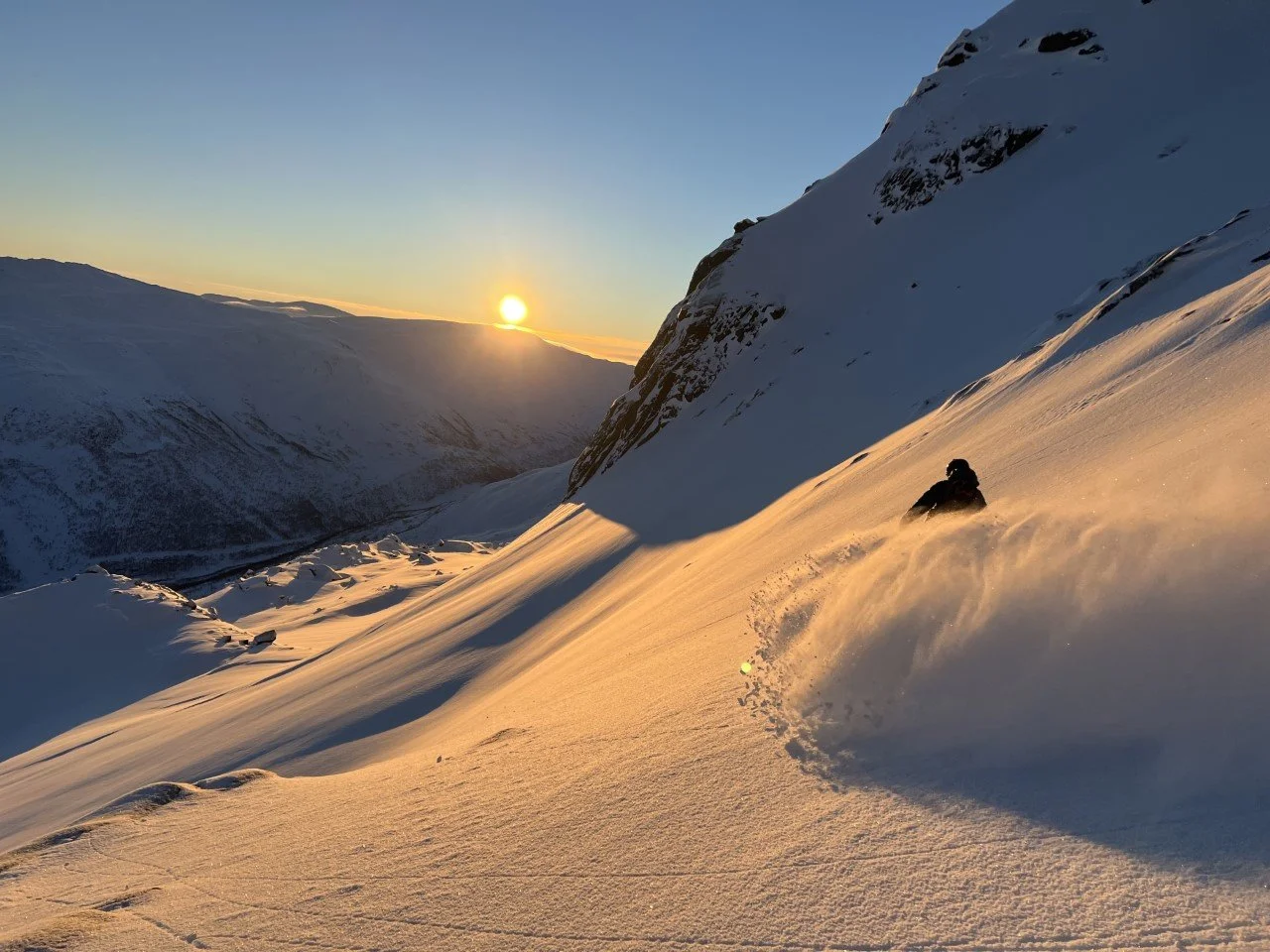En person kjører snøbrett ned en snødekt fjellskråning ved solnedgang. Snøen er nylig jordet, og landskapet er omgitt av fjell.