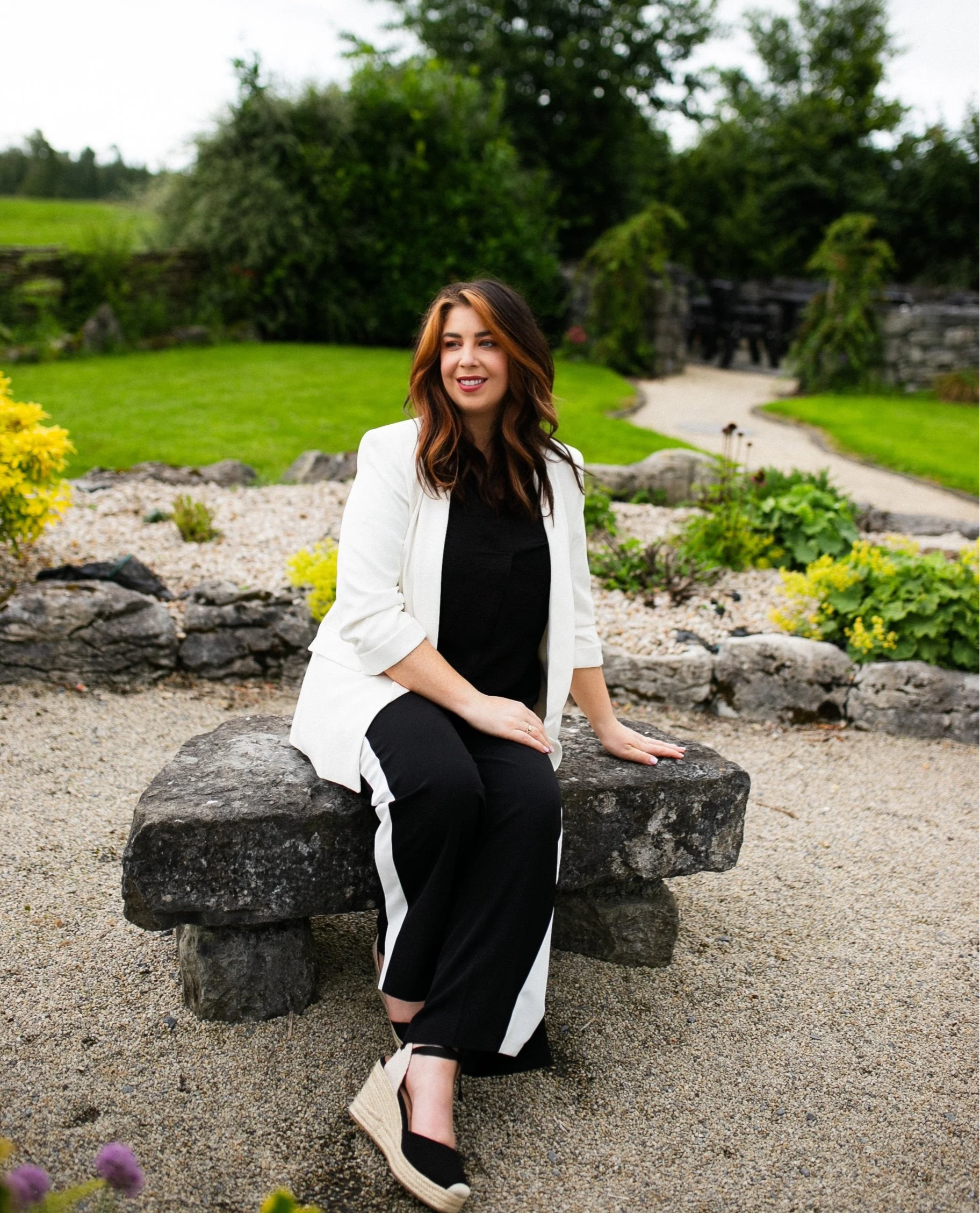 A young woman with brown hair and fair skin sitting on a large rock in a garden with green bushes and trees, wearing a white blazer, black top, black pants with white stripe, and beige wedge shoes, smiling and looking to her left.