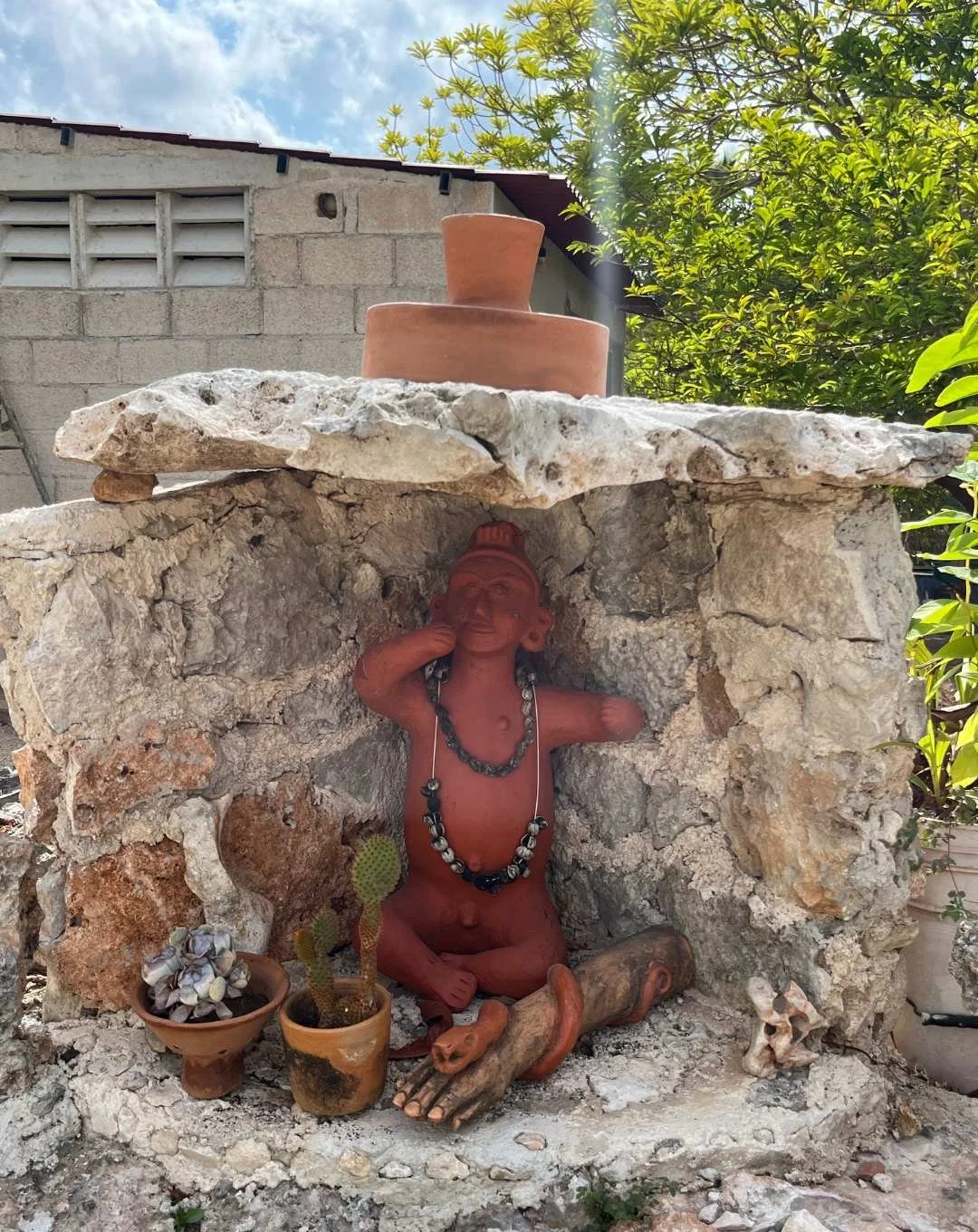 A mixed media outdoor sculpture of a seated, cross-legged figure with a necklace, inside a small rock shelter, underneath a large stone slab. There are two potted succulents and a small animal skull nearby, with a background of a building, trees, and