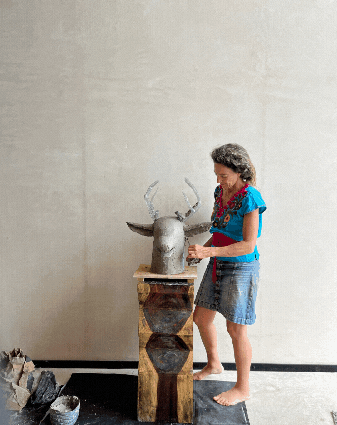A woman working on a sculpture of a deer's head with antlers, positioned on a wooden pedestal, in an art studio setting.