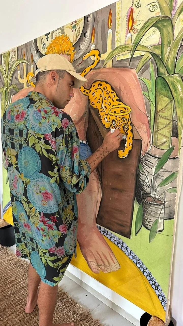 A person painting a colorful mural depicts a tropical scene with large plants, candles, and a leopard climbing on a rock formation.
