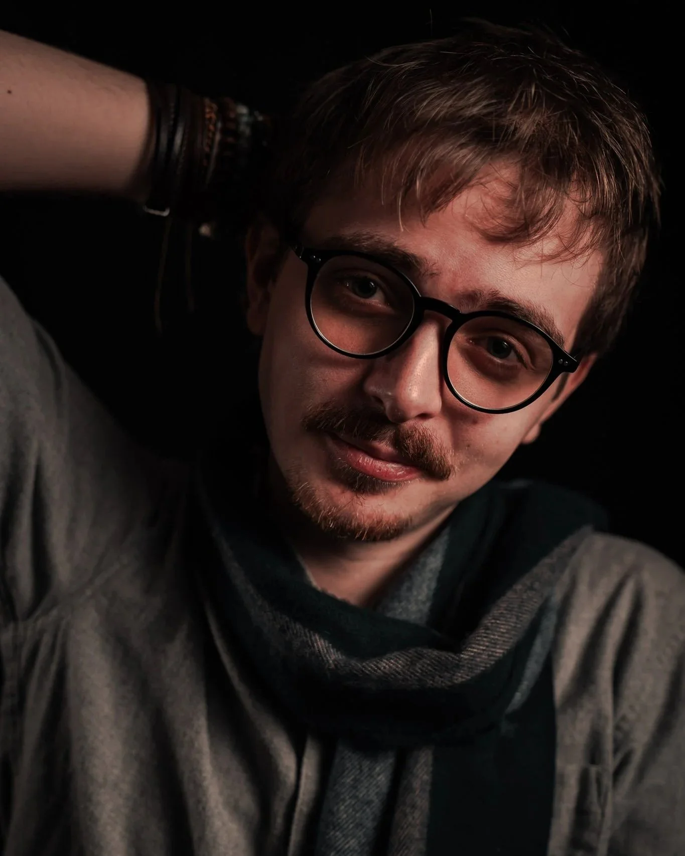 A young man with glasses, mustache, and beard, smiling, wearing a gray shirt and dark scarf, with his right hand behind his head against a black background.
