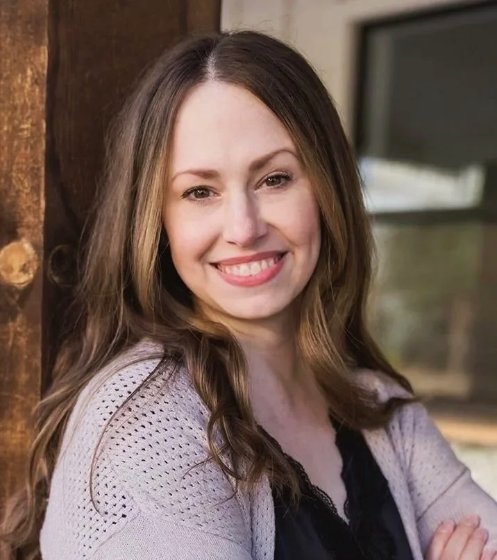 A young woman with long brown hair and a big smile, wearing a light-colored cardigan over a black top, standing outside near a wooden structure.