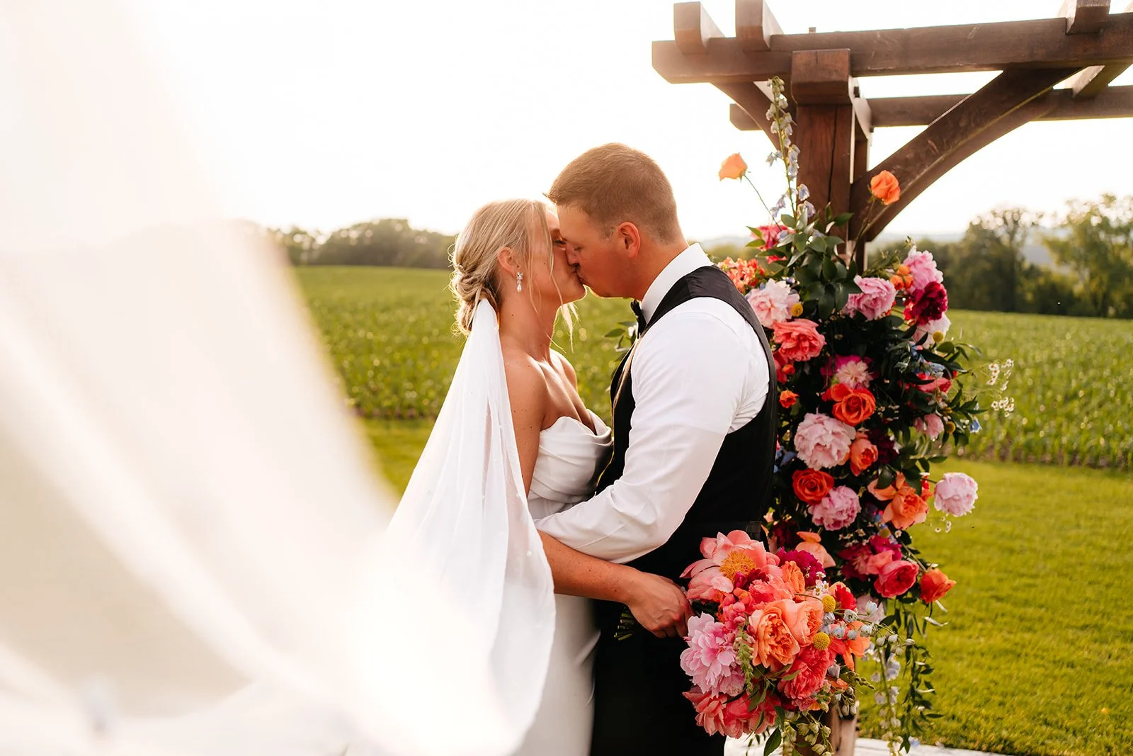 Bride and groom kissing under a wooden arbor adorned with vibrant coral, pink, and orange flowers by Darling Block Floral in Rochester, MN.