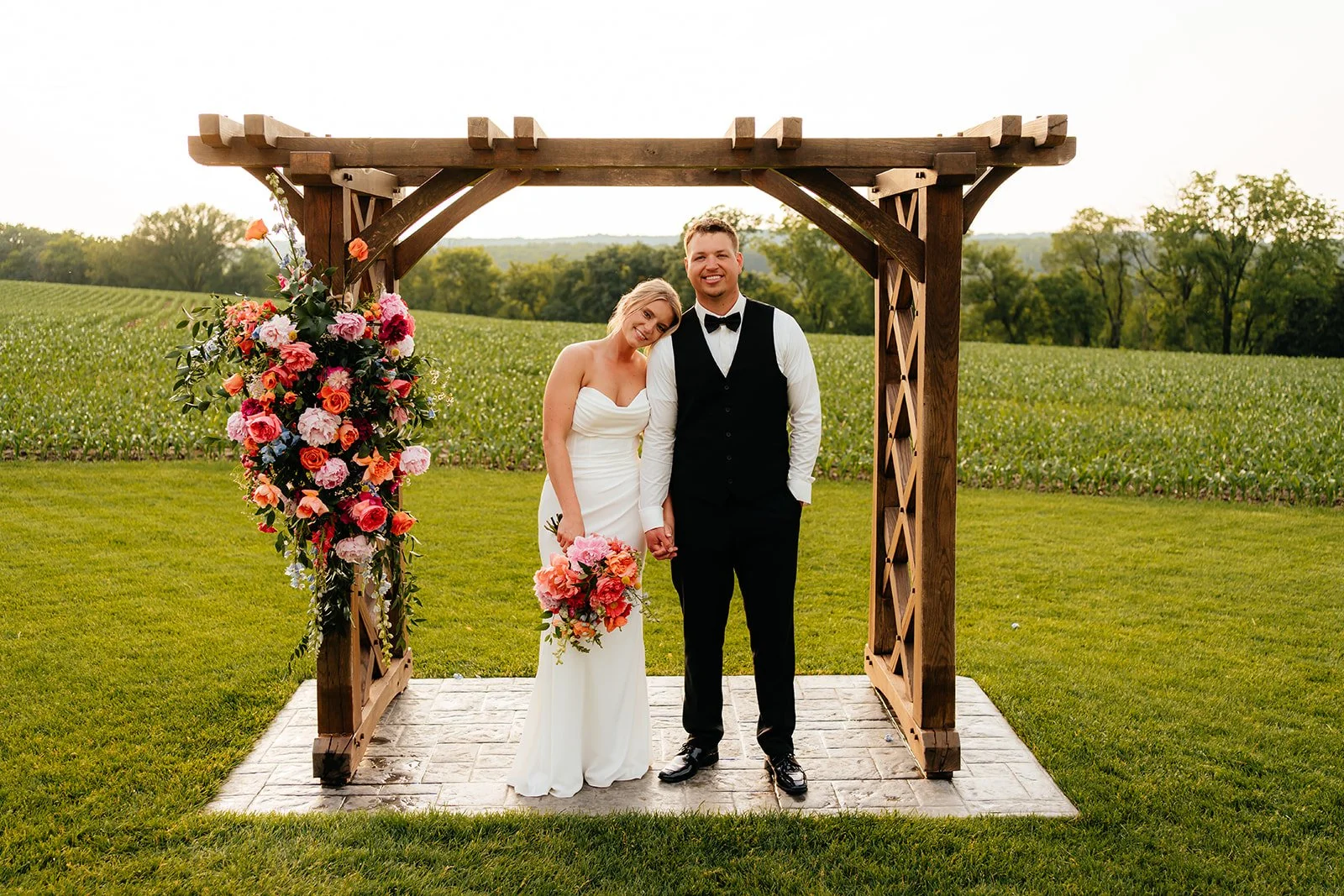 Bride and groom under a wooden arbor adorned with vibrant coral, pink, and orange flowers by Darling Block Floral in Rochester, MN.