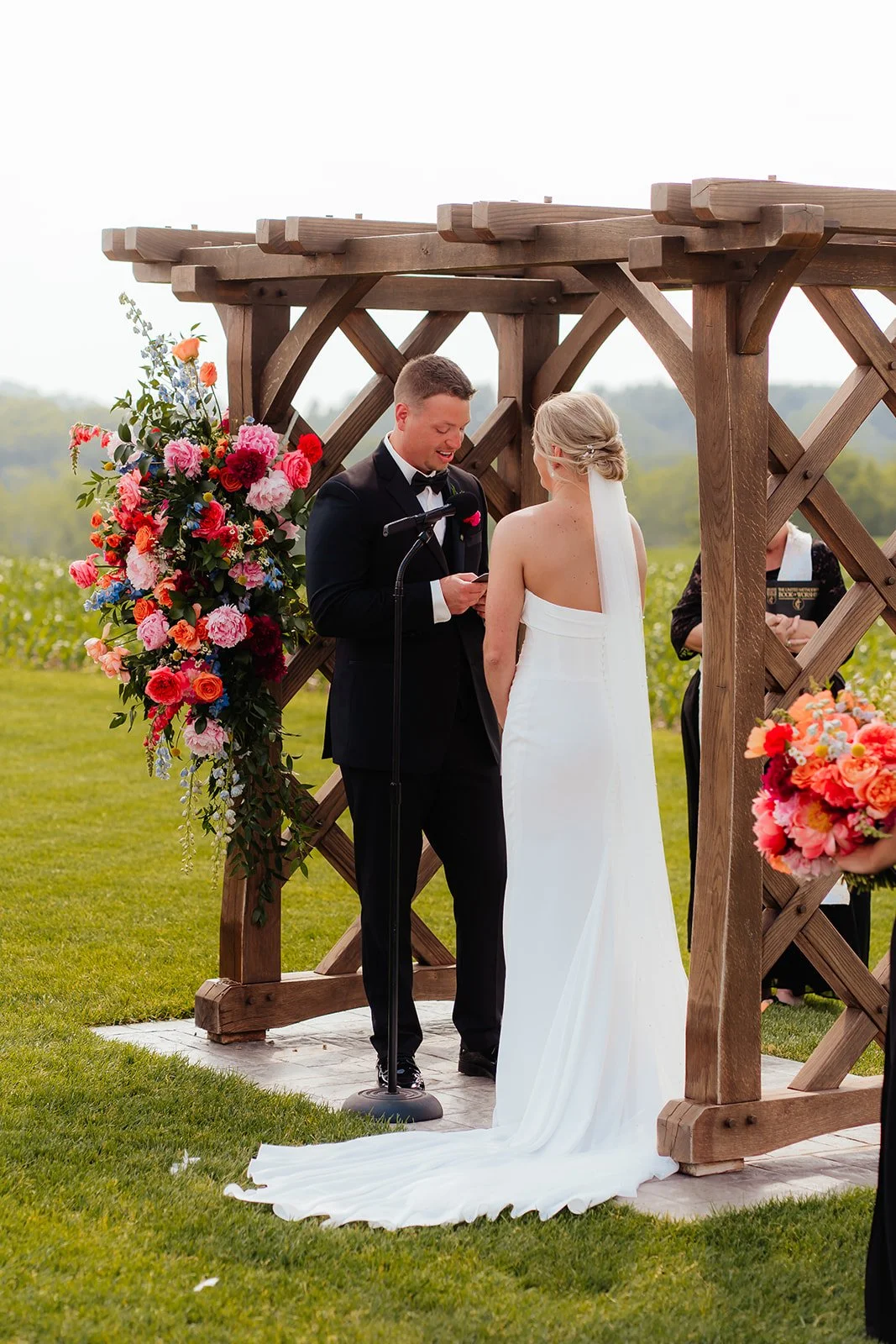 Bride and groom under a wooden arbor adorned with vibrant coral, pink, and orange flowers by Darling Block Floral in Rochester, MN.