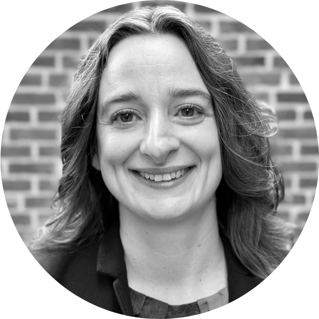 Black and white photo of a woman with shoulder-length wavy hair, wearing a blazer, smiling in front of a modern building.