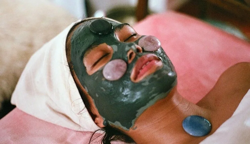 Person lying down with a facial mask, eye patches, and a jade roller on their face, resting on a pillow.
