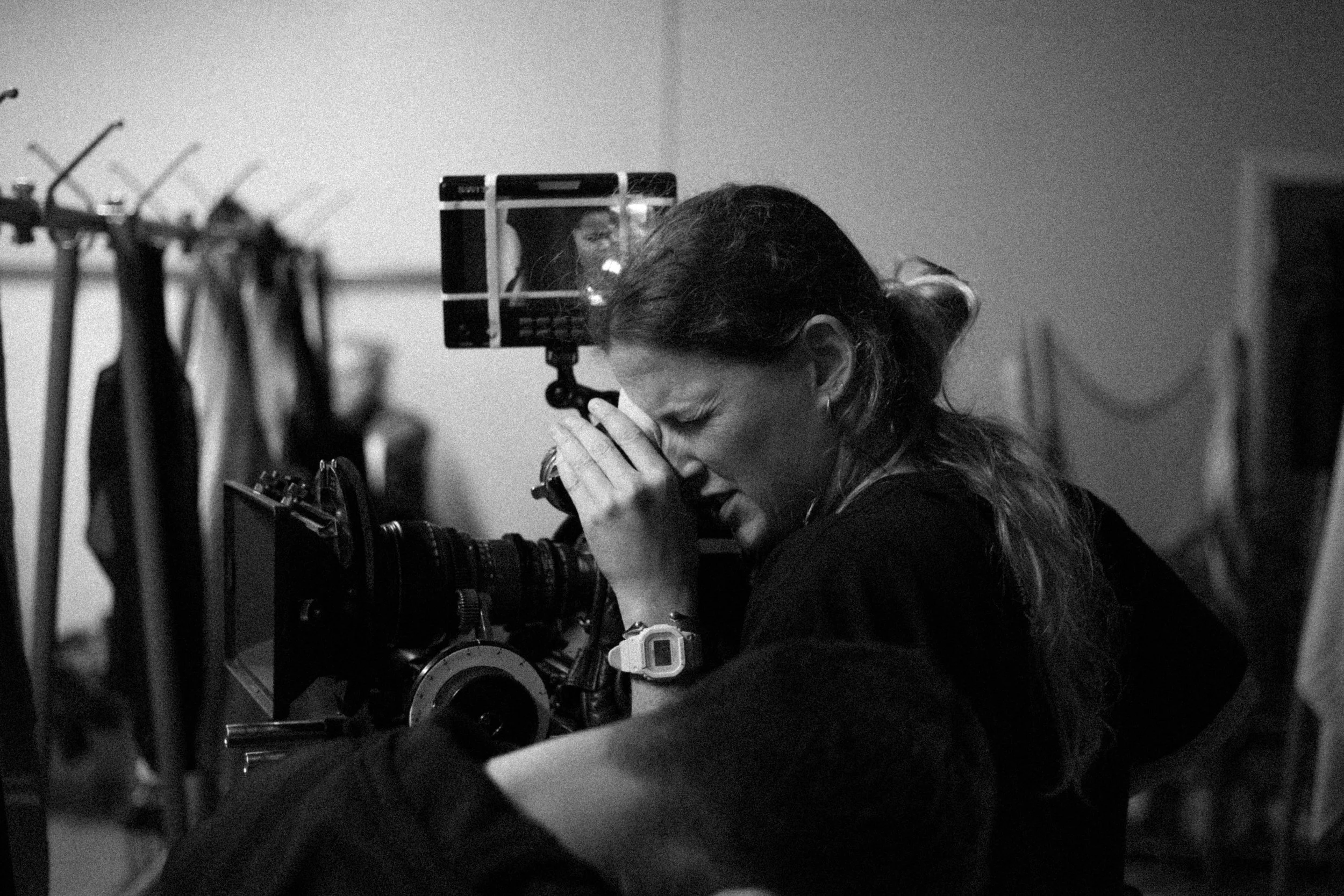 A woman operating a professional film camera, focusing through the viewfinder in a black and white photo.