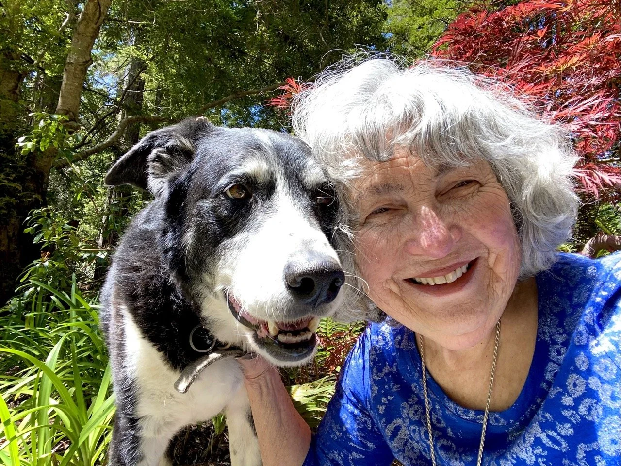 An elderly woman with white, curly hair smiling next to a black and white dog outdoors among trees and plants.