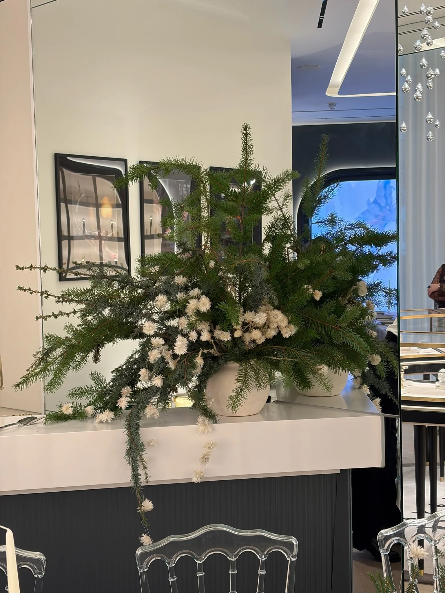 A large floral arrangement with green pine branches and white flowers in a white container on a white table in a modern interior with framed pictures and decorative lighting.