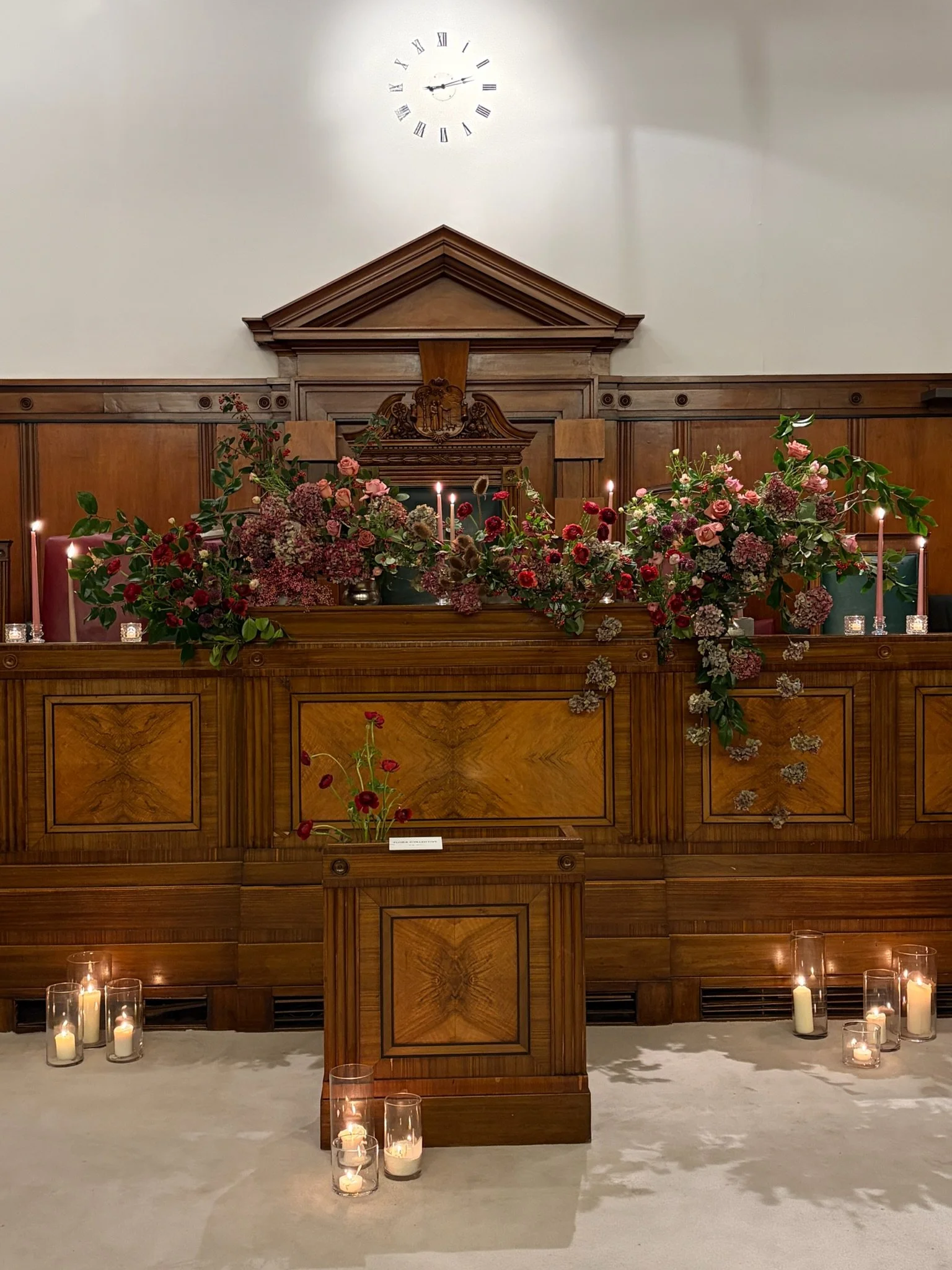 Indoor setting with a wooden altar decorated with red, pink, and purple flowers, lit candles, and a clock on the wall showing 2:15.