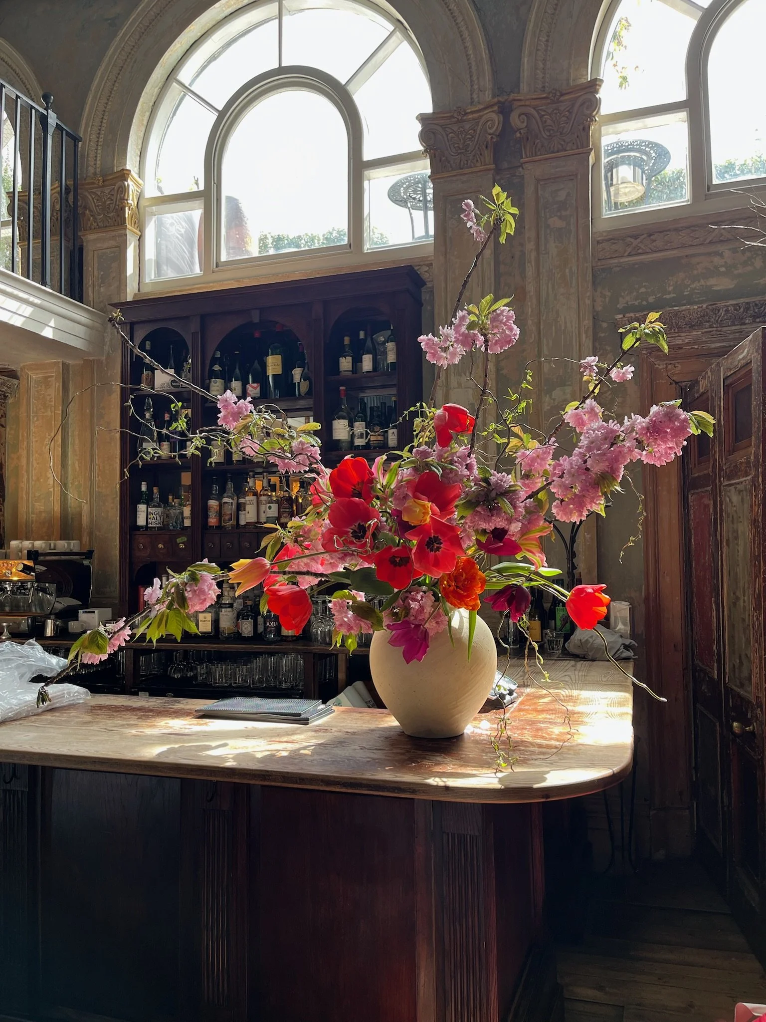 Large floral arrangement of pink, red, orange, and purple flowers in a cream-colored vase on a wooden bar or counter inside a well-lit, ornate room with arched windows and a dark wood bar with bottles of alcohol.