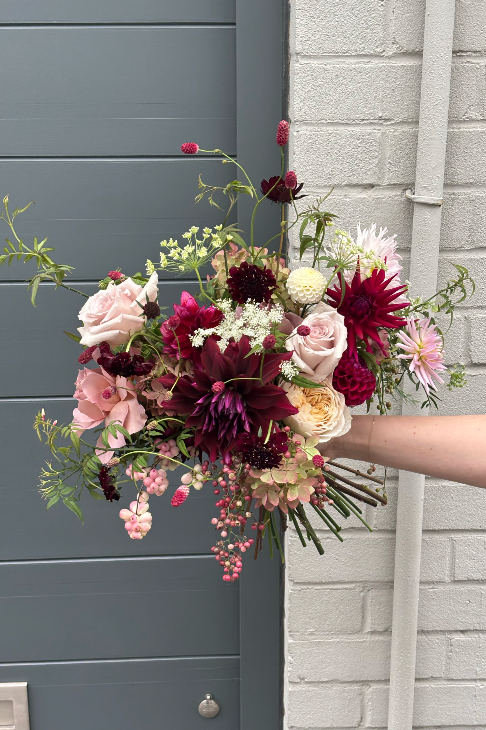 A hand holding a bouquet of mixed flowers including roses, dahlias, and berries, in front of a gray door and a white brick wall.