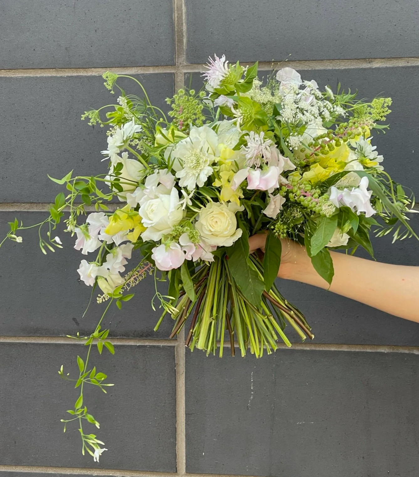 Hand holding a bouquet of white and pale pink flowers against a gray tile wall.