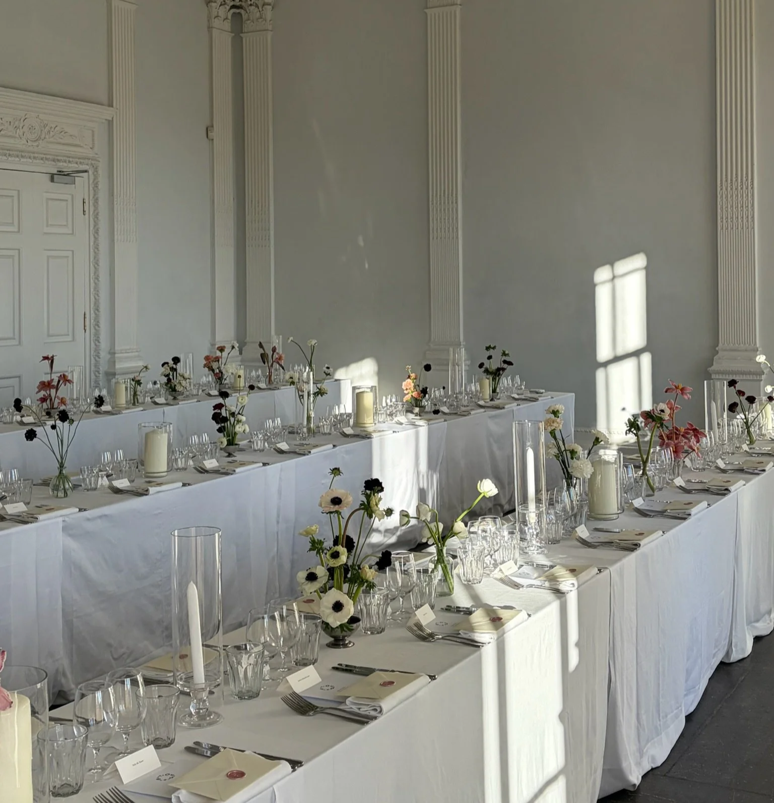 Elegant banquet table setup with white tablecloths, vases filled with white, pink, and black flowers, white candles in glass holders, and neatly arranged plates, silverware, and glasses, illuminated by natural sunlight streaming through windows.