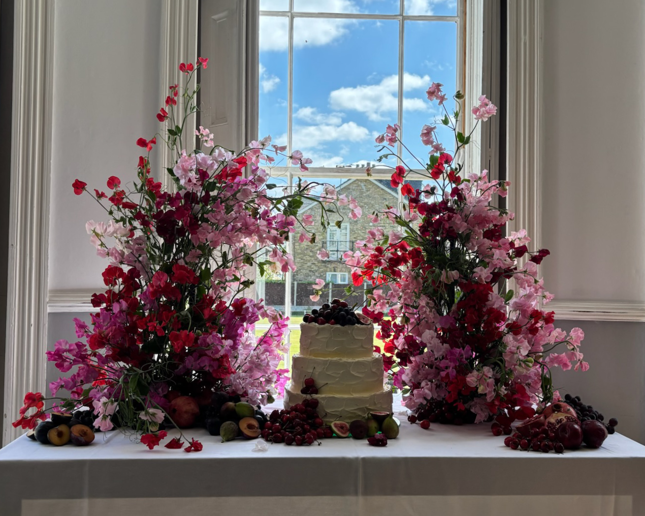 A wedding or celebration cake on a white table, decorated with dark berries, surrounded by large pink and red floral arrangements, with an open window showing blue sky and clouds in the background.