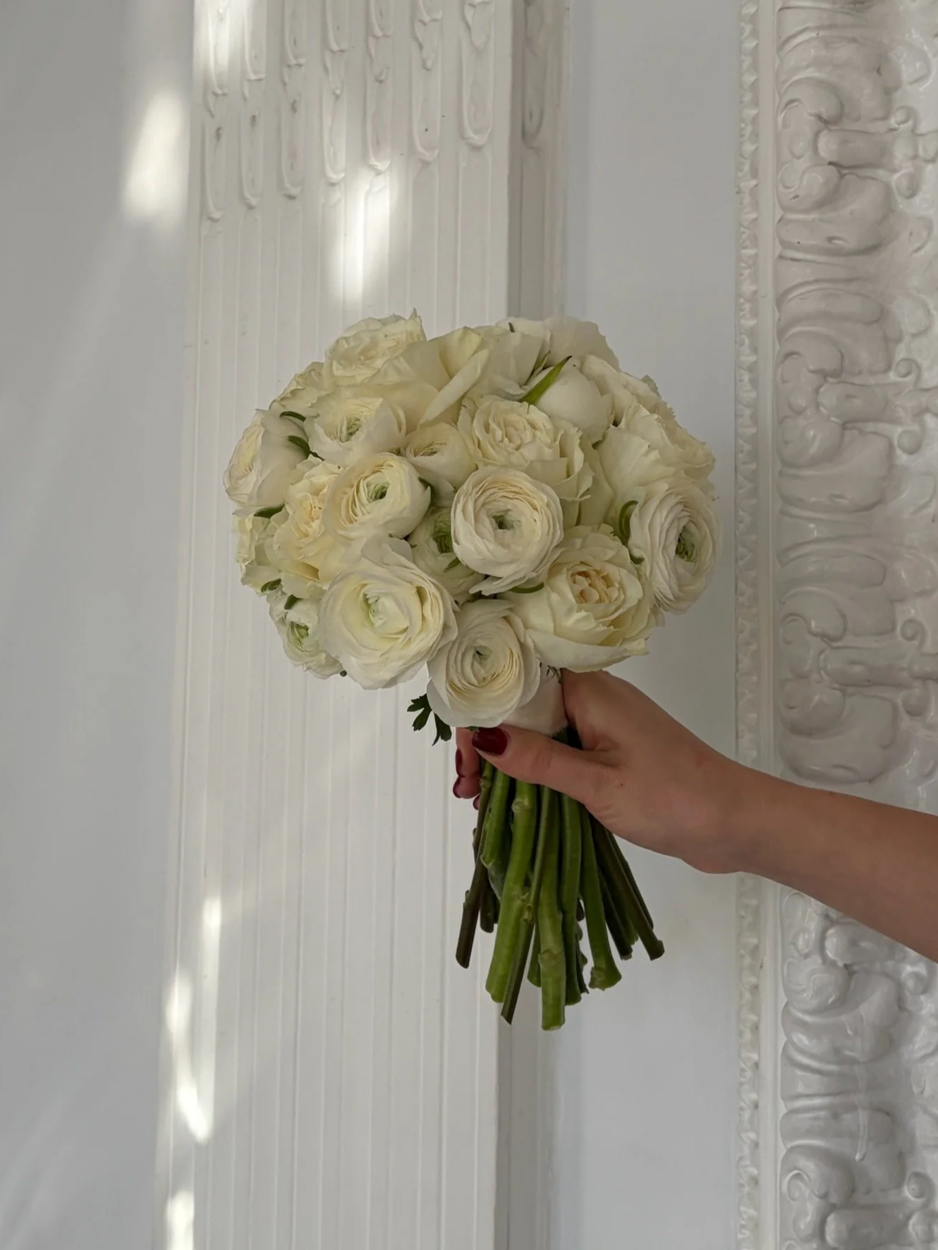A hand holding a bouquet of white flowers, including roses and ranunculus, against a white ornate background.