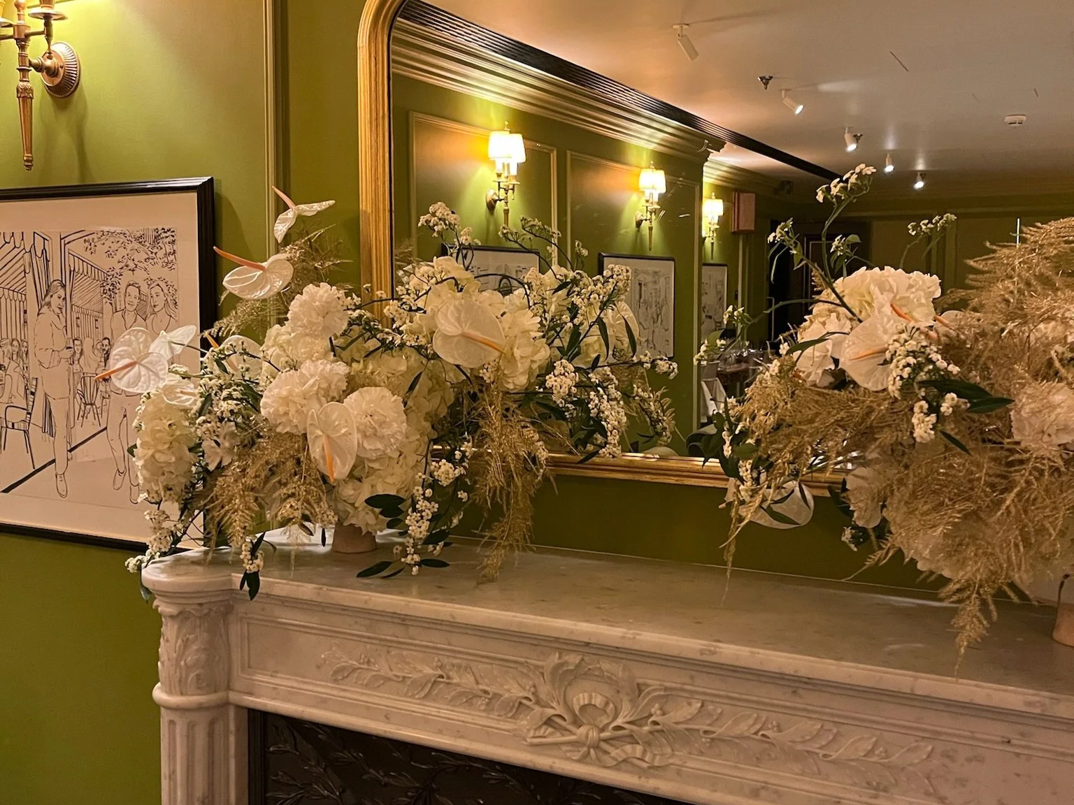 Decorative floral arrangements with white flowers, including orchids, on a marble fireplace mantel in a warmly lit room with green walls and framed artwork.