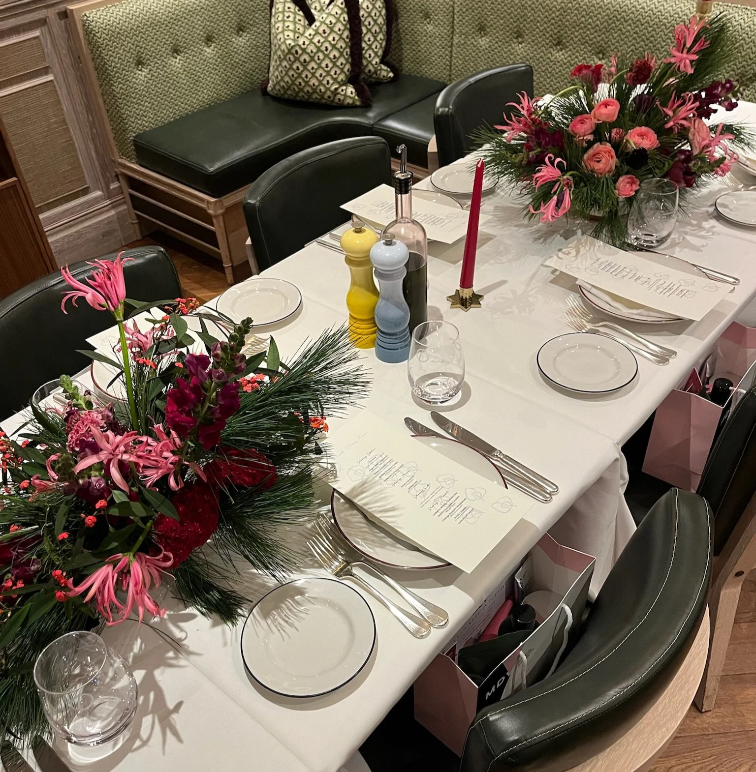 A dining table set for a formal gathering with floral centerpieces, menus, glassware, utensils, and decorative salt and pepper shakers, along with a pink candle, in a room with green upholstered seating.