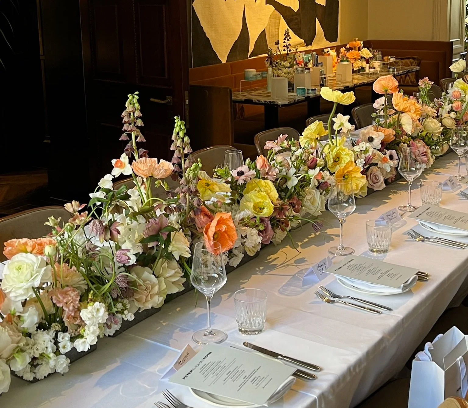 A long dining table decorated with a vibrant arrangement of pink, purple, white, and yellow flowers. The table is set with white plates, silverware, wine glasses, and menus, ready for a formal meal.