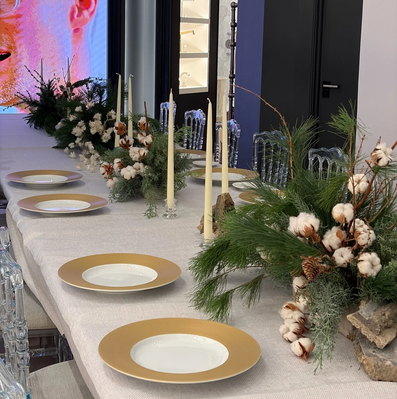 Decorated dining table with white tablecloth, gold-rimmed plates, tall candles, greenery, cotton branches, pine cones, and a large screen in the background.