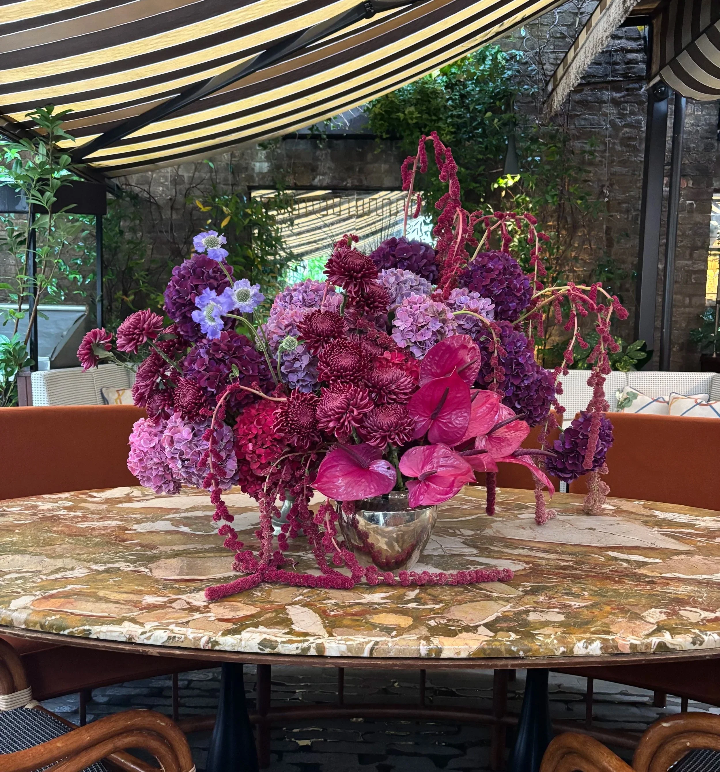 A vibrant floral centerpiece with purple, pink, and lavender flowers in a silver vase, placed on a marble table inside a decorated indoor space with striped awnings and greenery.
