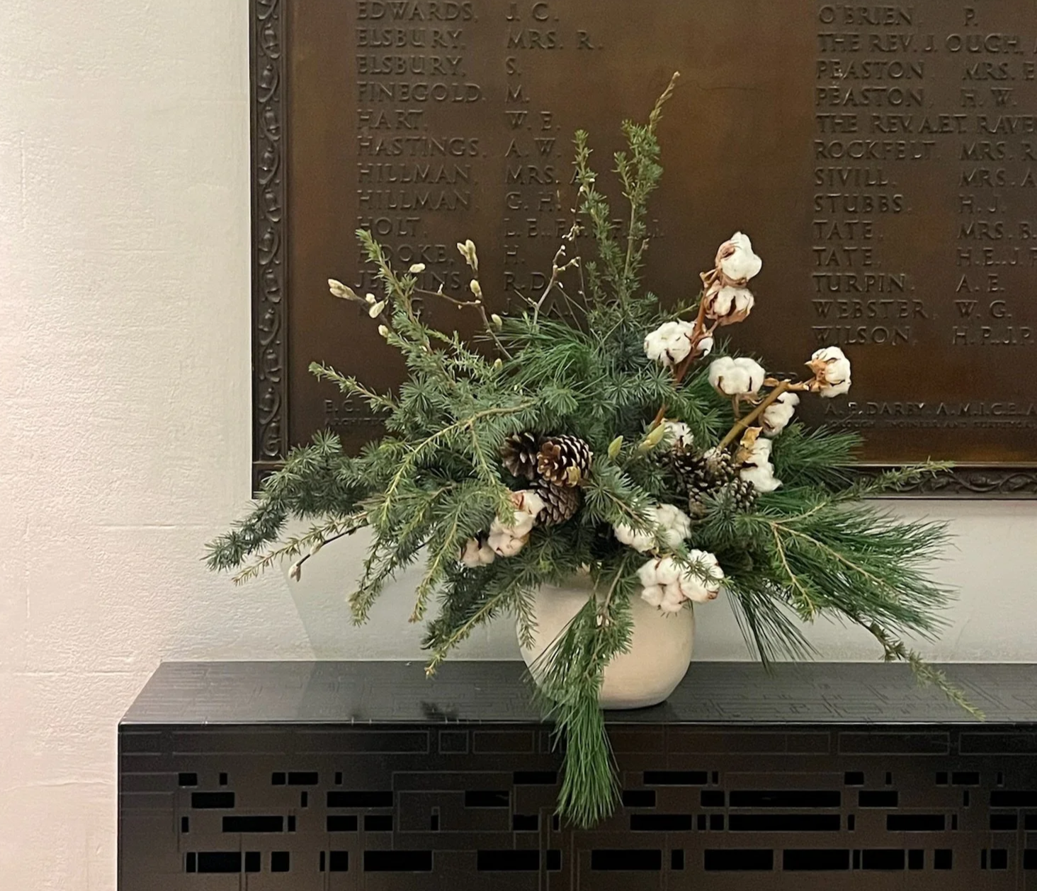 A floral arrangement in a white vase with cotton stems, pine branches, and pinecones placed on a black decorative table in front of a bronze memorial plaque with inscriptions of names.