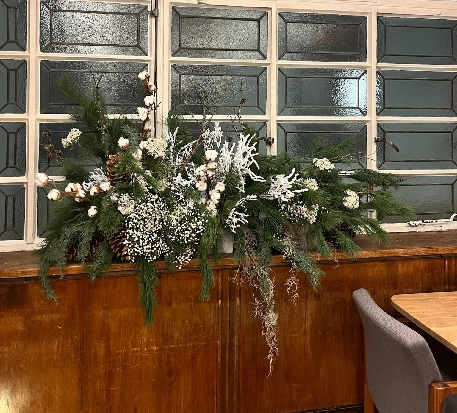 A large winter floral arrangement with pine branches, cotton, baby's breath, and frosted decorative elements on a wooden cabinet in front of stained glass windows.