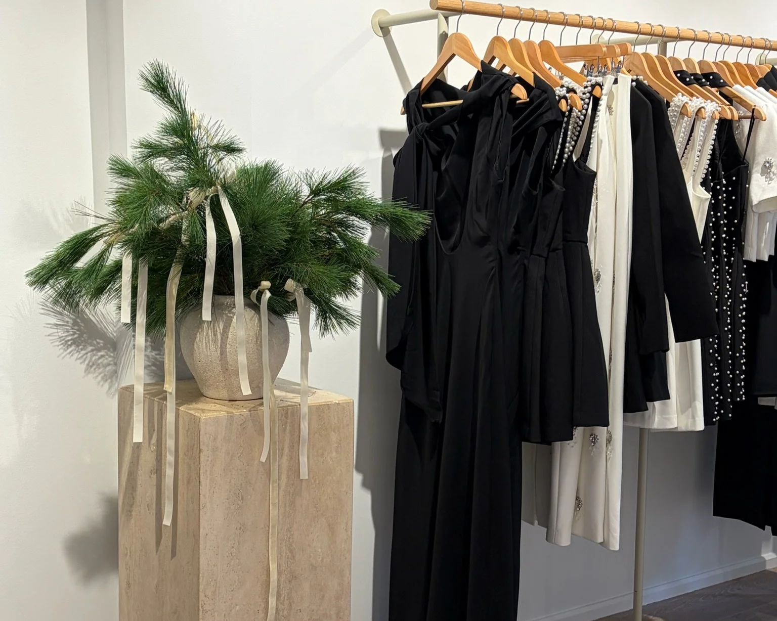 Clothing rack with black and white dresses, beside a beige pedestal with a potted pine tree decorated with cream ribbons for Christmas.