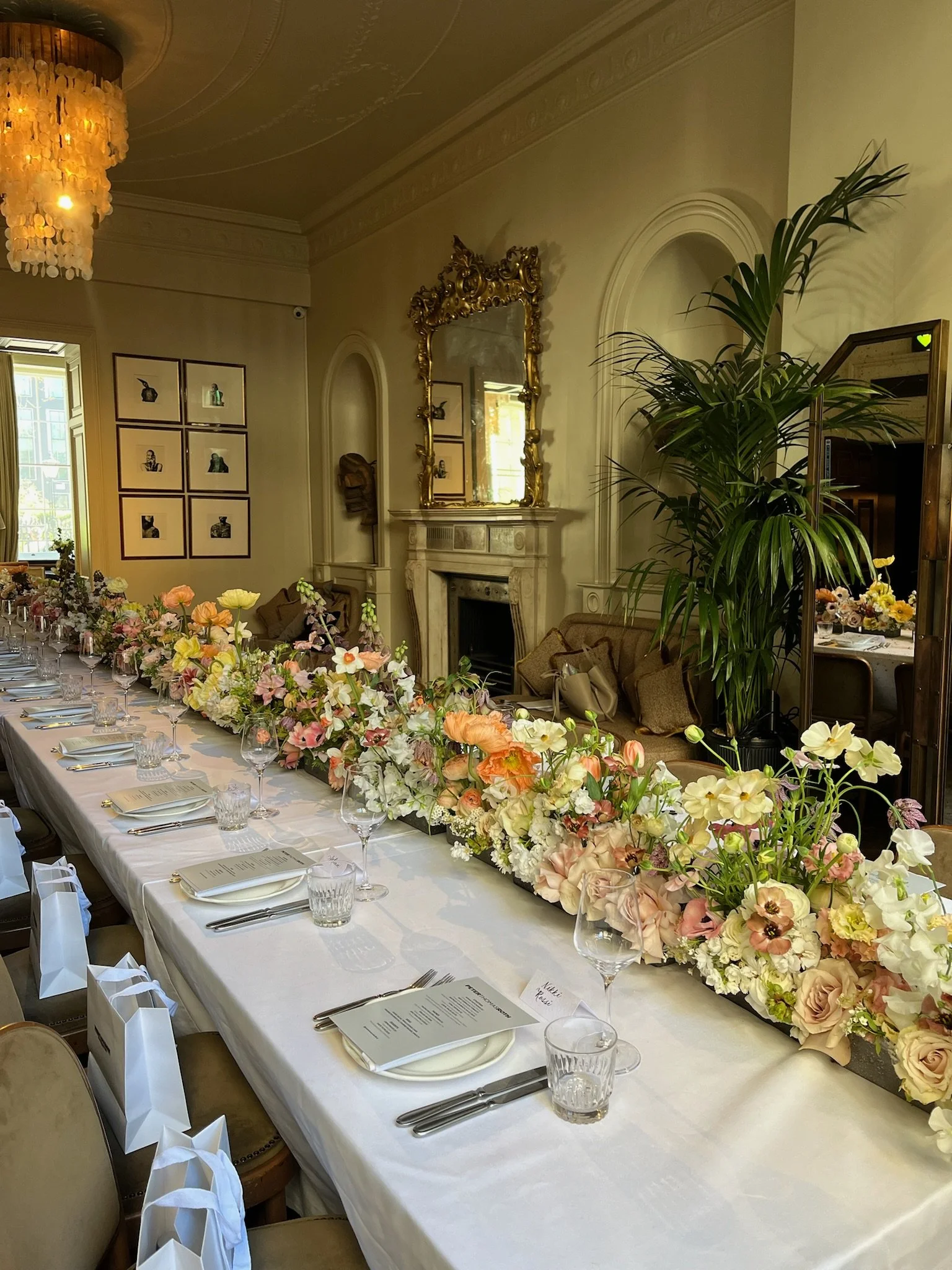 A long dining table decorated with an abundance of pastel-colored flowers and place settings, inside an elegant room with a gold-framed mirror, a marble fireplace, and a large green plant.