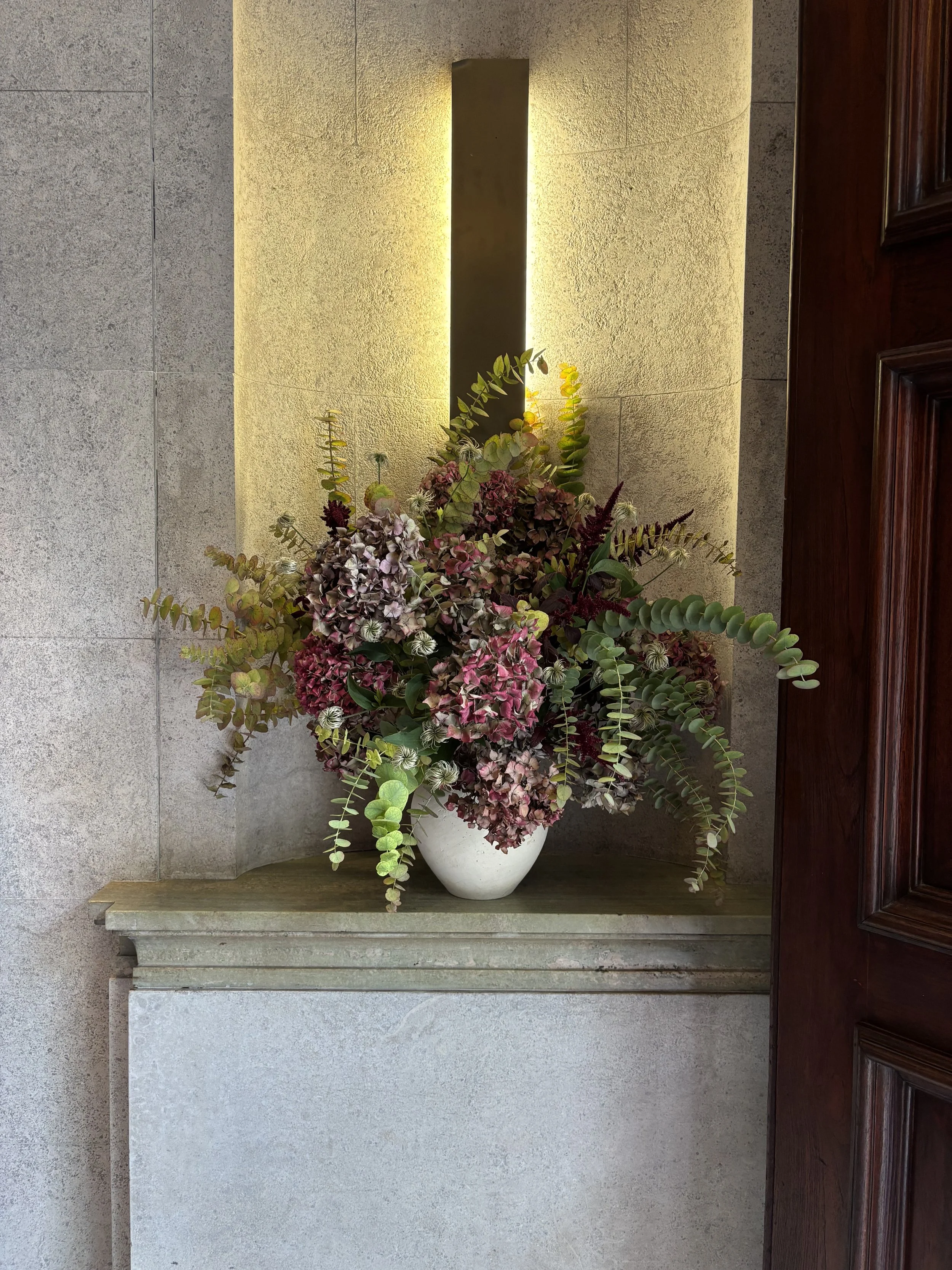 A floral arrangement with pink, purple, and white flowers and green foliage in a white vase on a stone pedestal against a wall with lighting above.