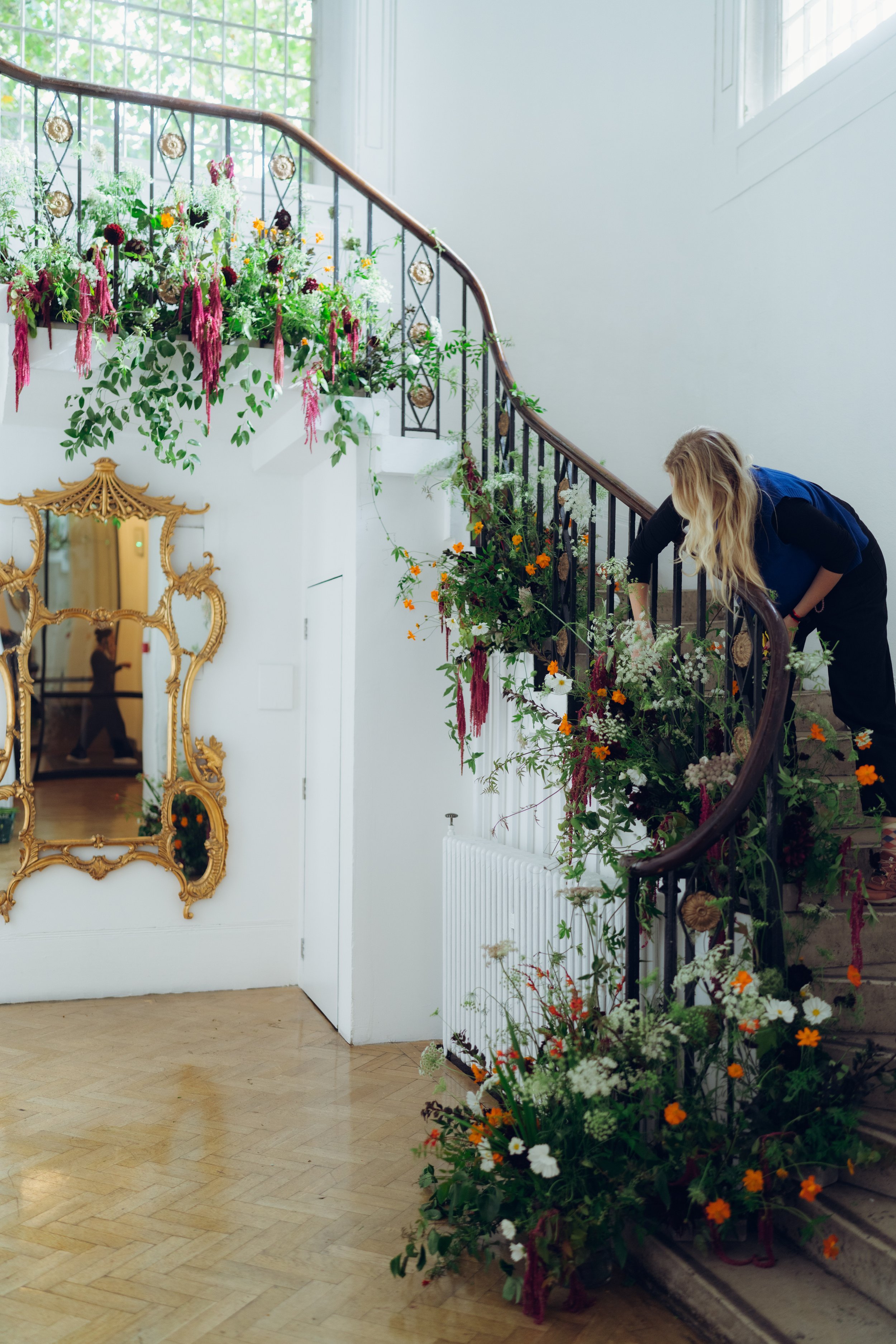 A woman is decorating a staircase with flowers and greenery in a bright, elegant interior with a large mirror and white walls.